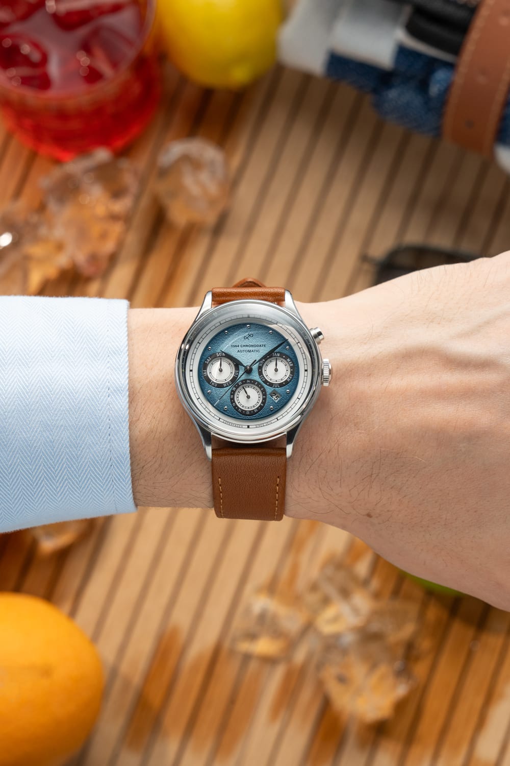 Wrist wearing the 1564 Chronodate Automatic by AV86 featuring a blue chronograph dial with three subdials, sapphire crystal, and a classic brown leather strap in a relaxed, warm-toned setting.