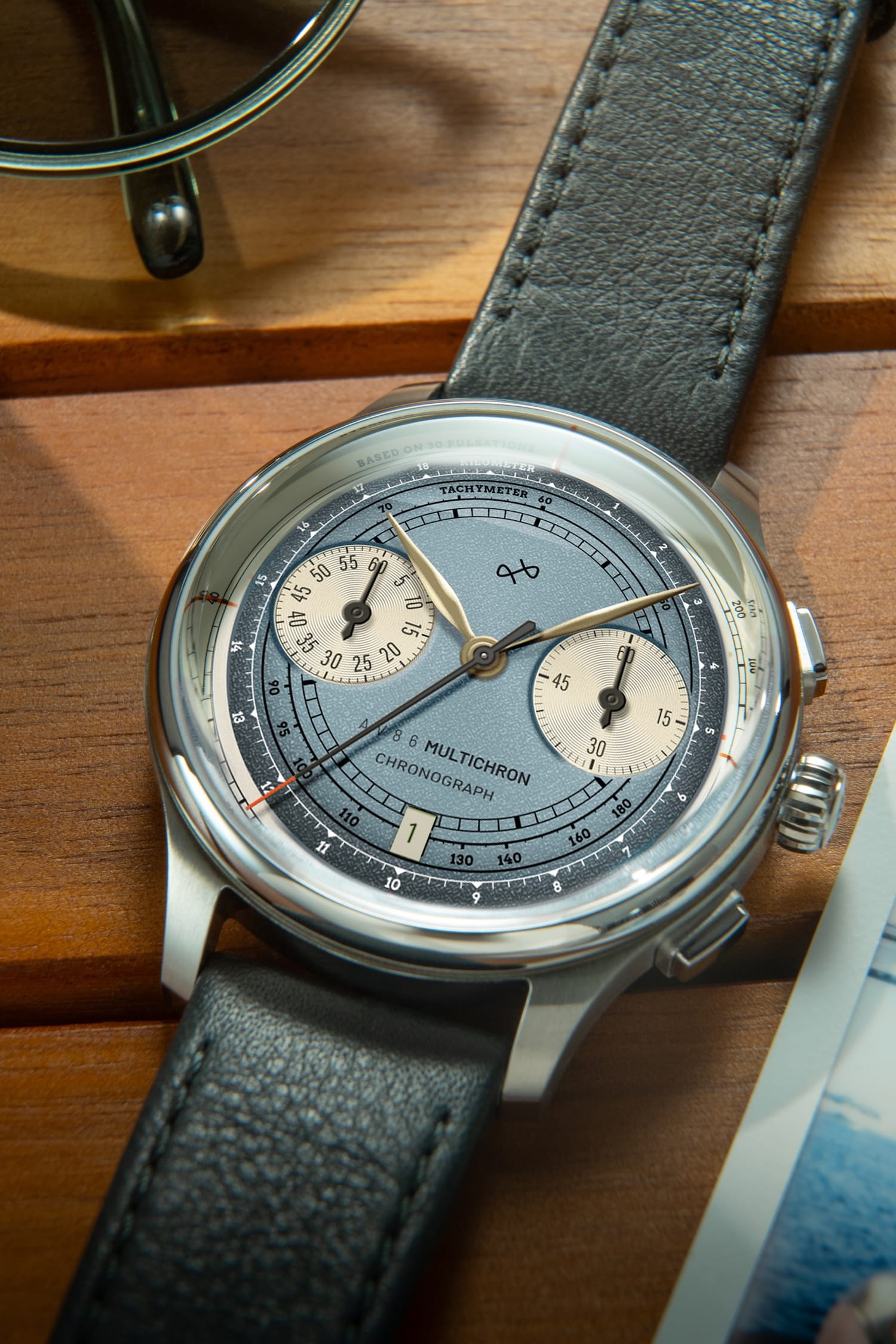 1934 Multichron, Steel / Sky Blue by AV86 featuring a vintage-inspired chronograph with a sapphire crystal, detailed tachymeter scale, and textured leather strap resting on a wooden surface.