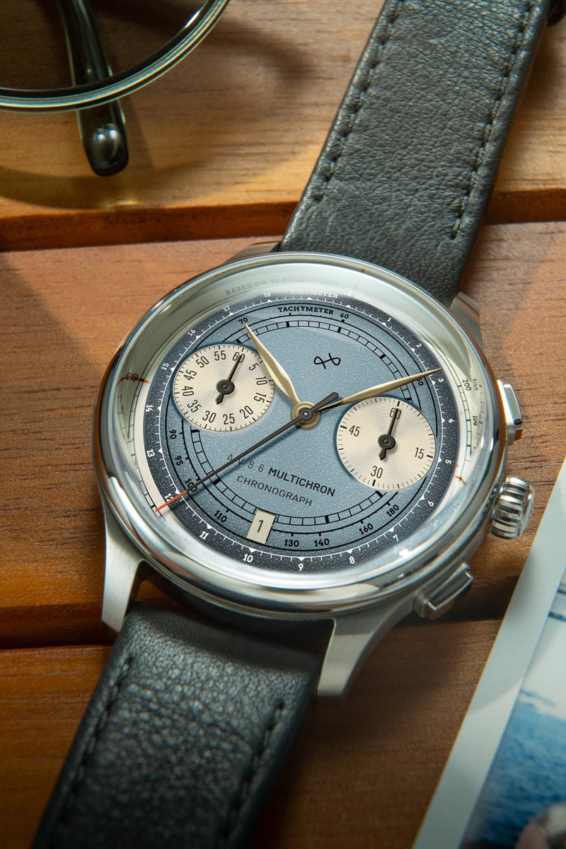 1934 Multichron, Steel / Sky Blue by AV86 featuring a vintage-inspired chronograph with a sapphire crystal, detailed tachymeter scale, and textured leather strap resting on a wooden surface.
