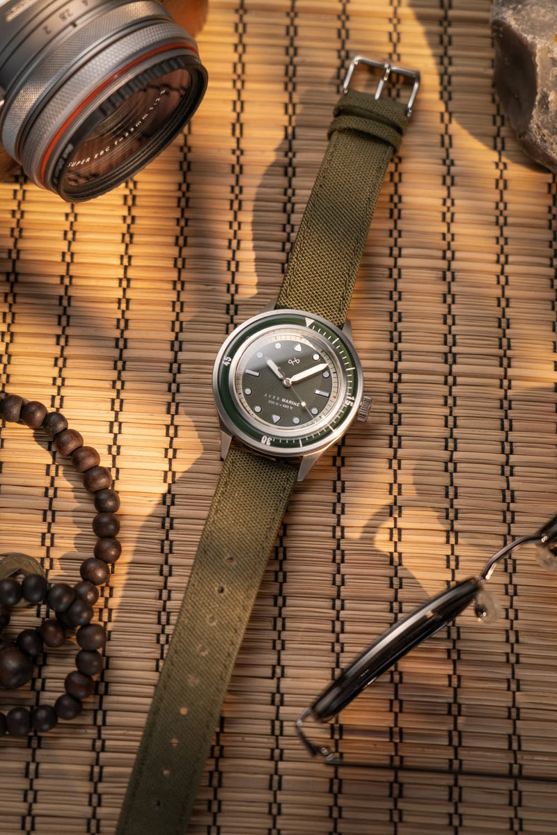 1956 Marine by AV86 vintage-inspired diver with sapphire crystal, compass bezel, and textured green fabric strap on woven surface