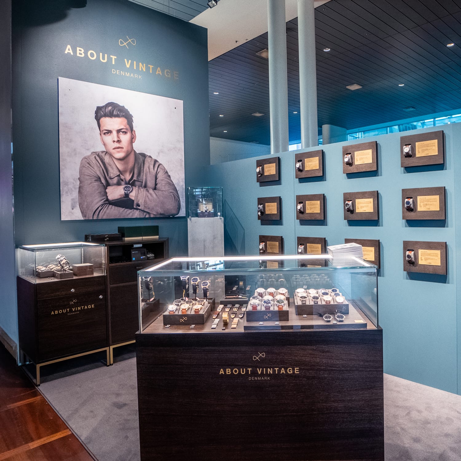 Showcase area featuring vintage-inspired wristwatches in glass cases with a large portrait and award plaques on blue walls background.