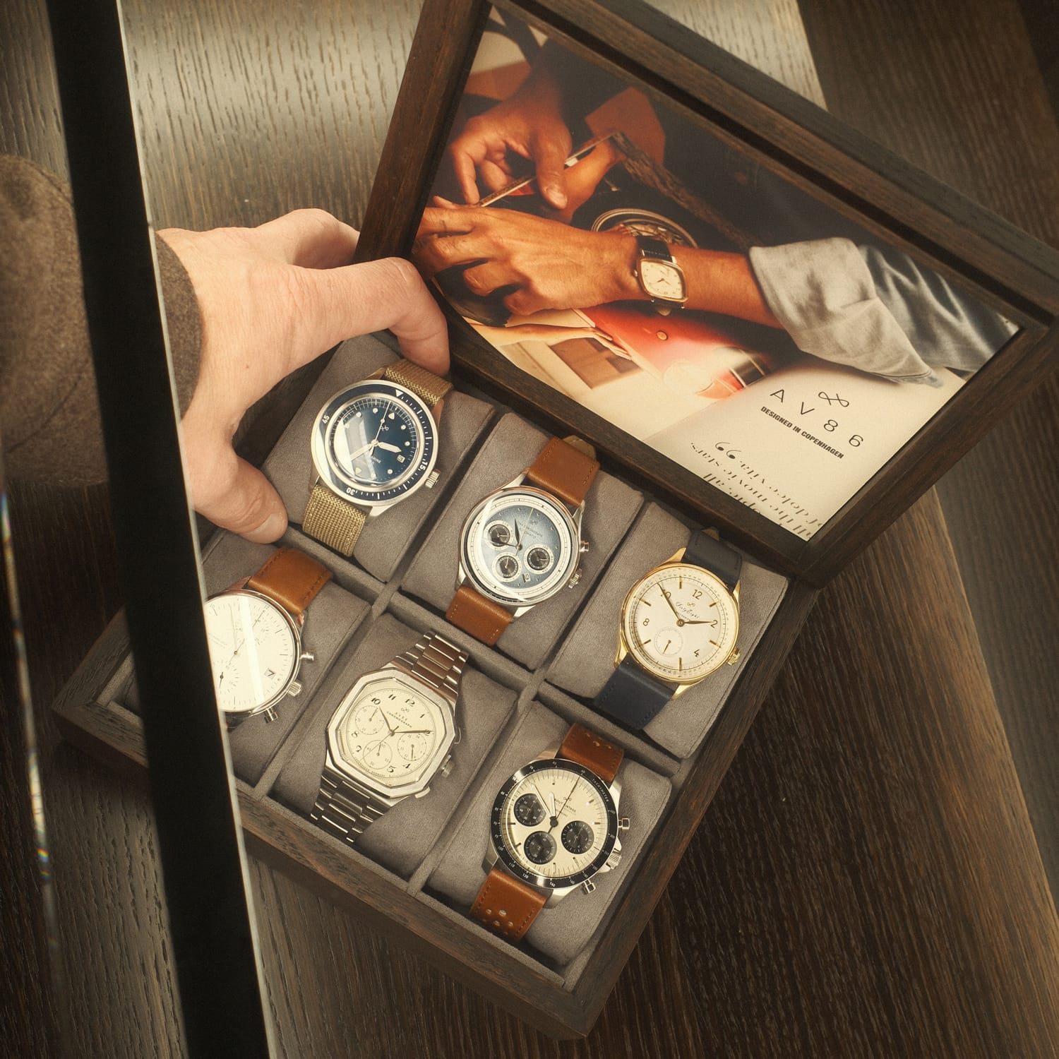 Wooden display box holding six wristwatches with various dial designs and straps, one watch being lifted by a hand.