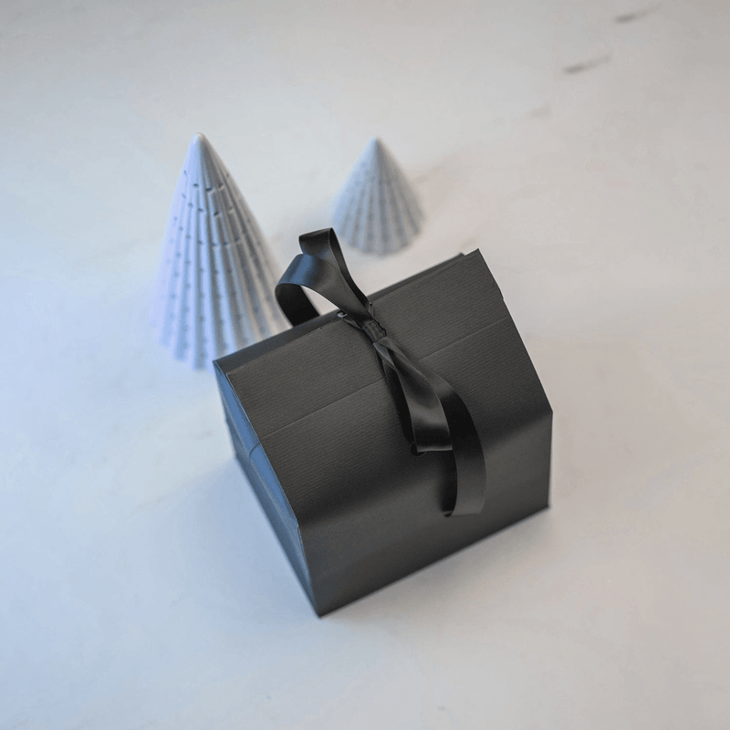Gift wrapping by AV86 featuring a sleek black box tied with a satin ribbon beside two minimalist paper cones on white surface.
