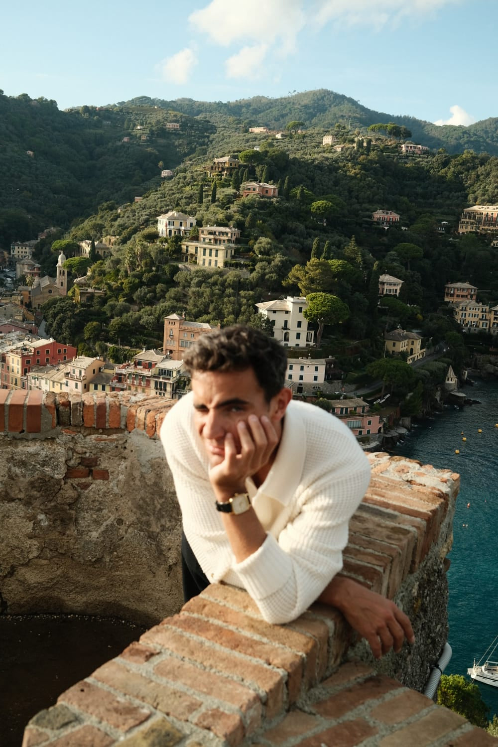 Man leaning on stone wall overlooking coastal hillside village, wearing cream knit sweater and wristwatch at sunset