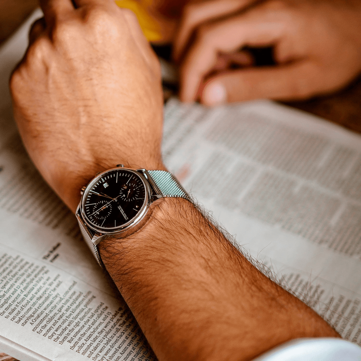 1844 Chronograph by AV86 with vintage-inspired dial, chronograph function, sapphire crystal, and mesh metal strap on wrist reading newspaper