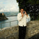 Man wearing 1954 Amalfi Mechanical, Steel / Off White by AV86, holding a camera near a scenic waterfront with stone railing.