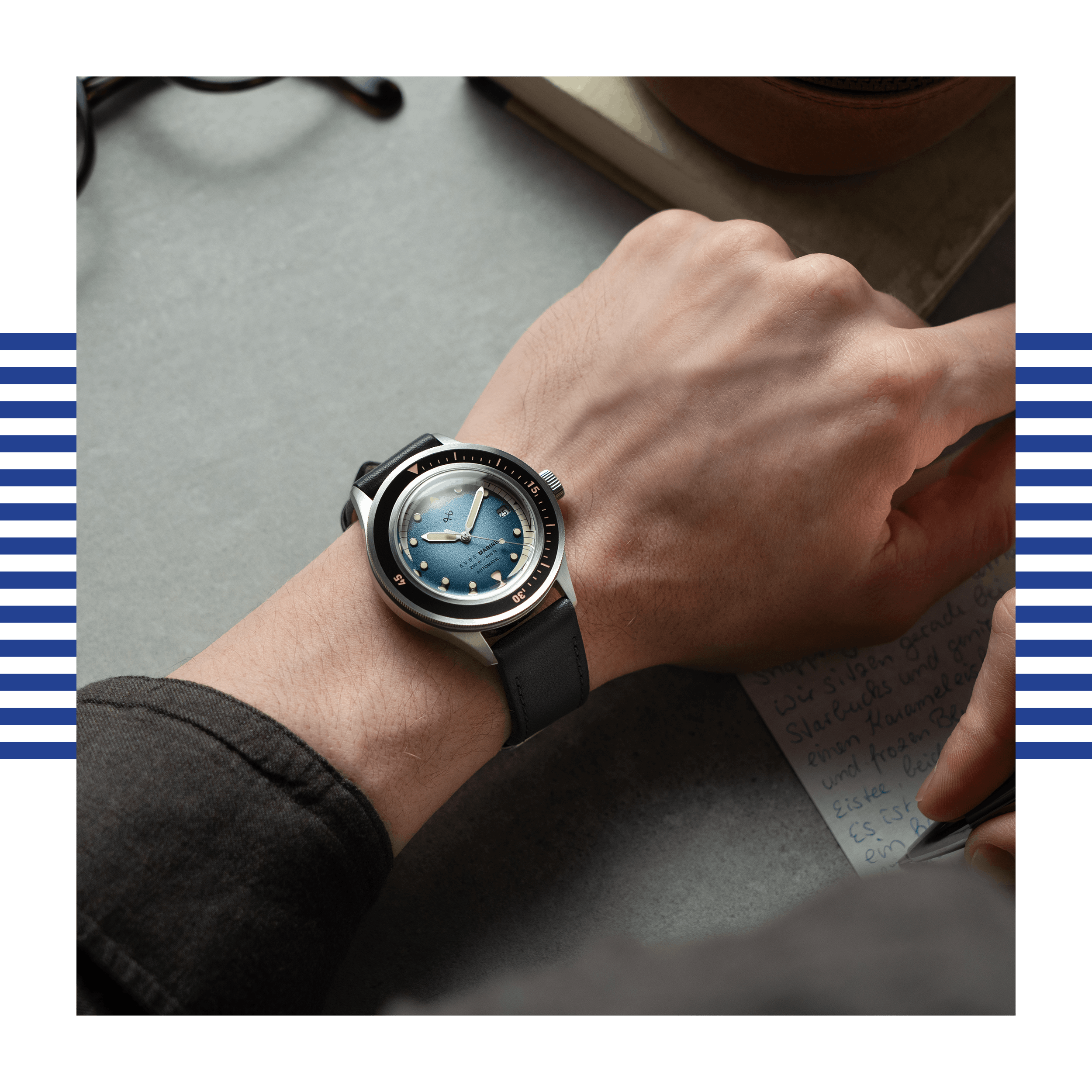 Wrist wearing a stainless steel watch with a blue gradient dial and black leather strap over a gray surface while writing.