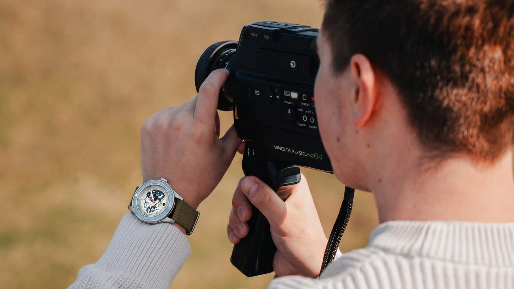 Man wearing wristwatch with olive green strap holds vintage film camera outdoors against blurred background.