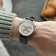 1564 Chronodate Automatic by AV86 displays a vintage-inspired automatic chronograph with a textured dial, triple subdials, polished stainless steel case, sapphire crystal, and black leather strap, worn in a cozy setting featuring soft knitwear and baked goods, embodying Nordic minimalism and Danish horological excellence.