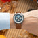 Wrist wearing the 1564 Chronodate Automatic by AV86 featuring a blue chronograph dial with three subdials, sapphire crystal, and a classic brown leather strap in a relaxed, warm-toned setting.