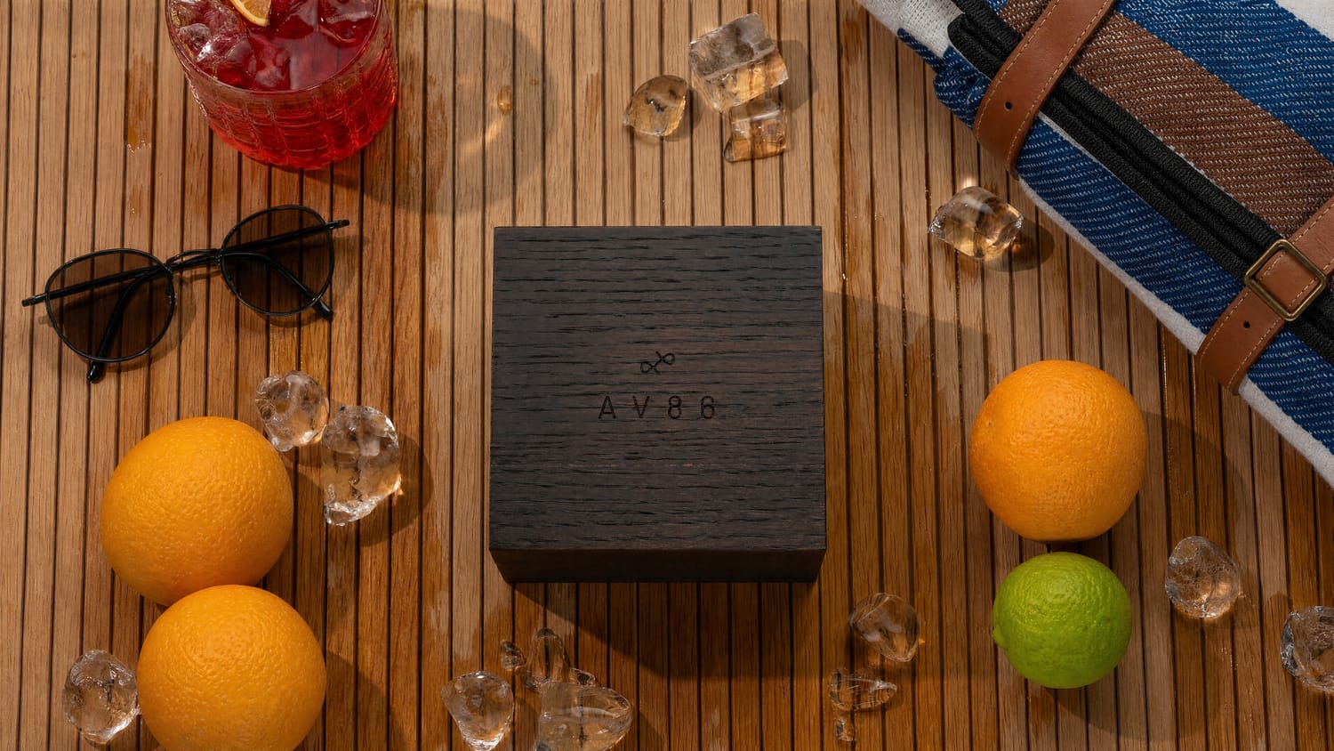 A minimalist AV86 wooden watch box set on a teak surface surrounded by fresh citrus fruits, sunglasses, a blue and brown striped fabric bag, scattered ice cubes, and a red cocktail evoking a refined Scandinavian summer lifestyle and complementing the 1564 Chronodate Automatic, Steel / Ice Blue & White packaging.