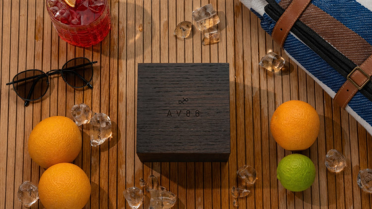 A minimalist AV86 wooden watch box set on a teak surface surrounded by fresh citrus fruits, sunglasses, a blue and brown striped fabric bag, scattered ice cubes, and a red cocktail evoking a refined Scandinavian summer lifestyle and complementing the 1564 Chronodate Automatic, Steel / Ice Blue & White packaging.