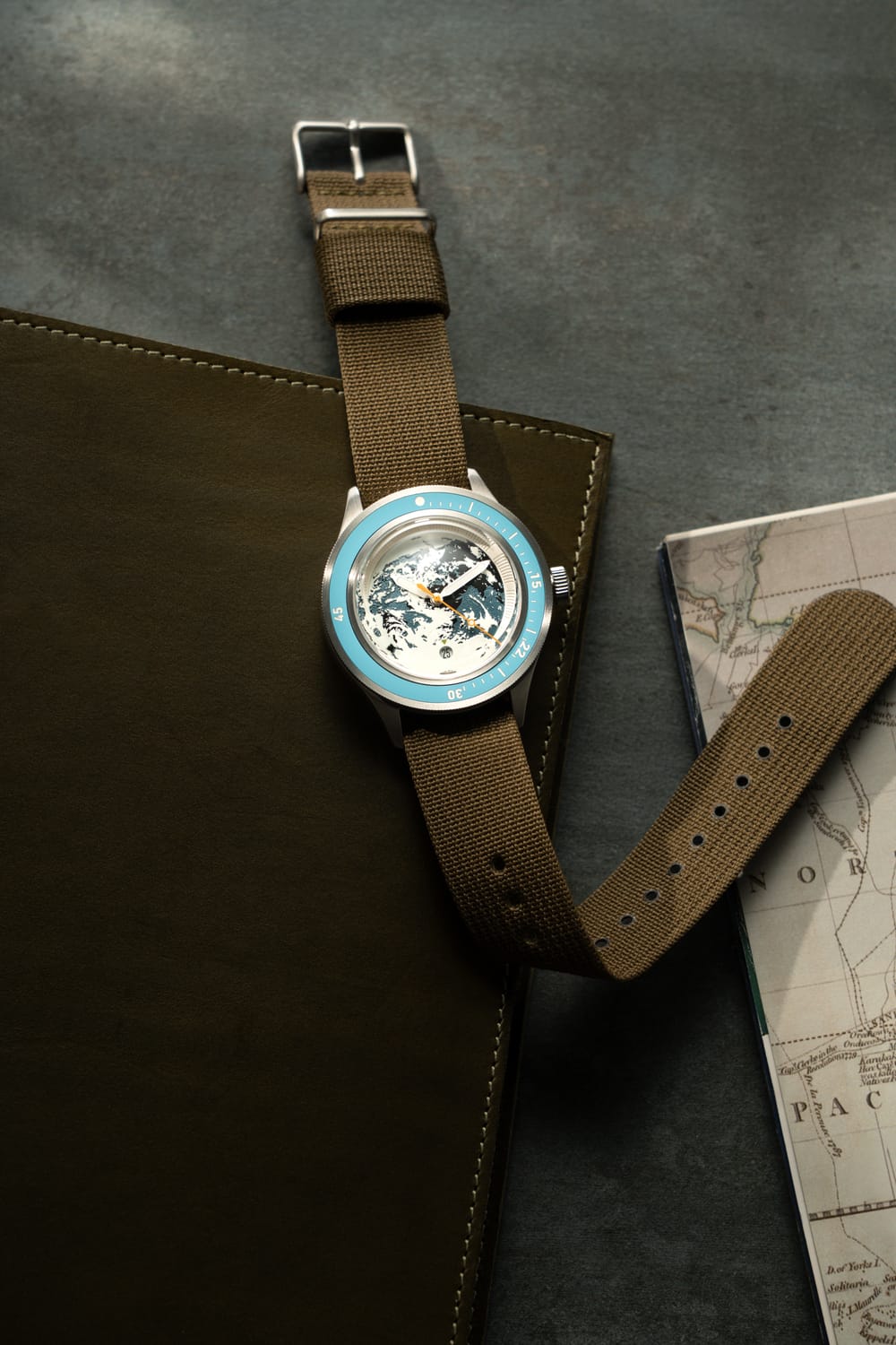 Earthrise WWF Partnership, Automatic / 2026 Edition by AV86 with a blue bezel and brown fabric strap, beside map and leather folio