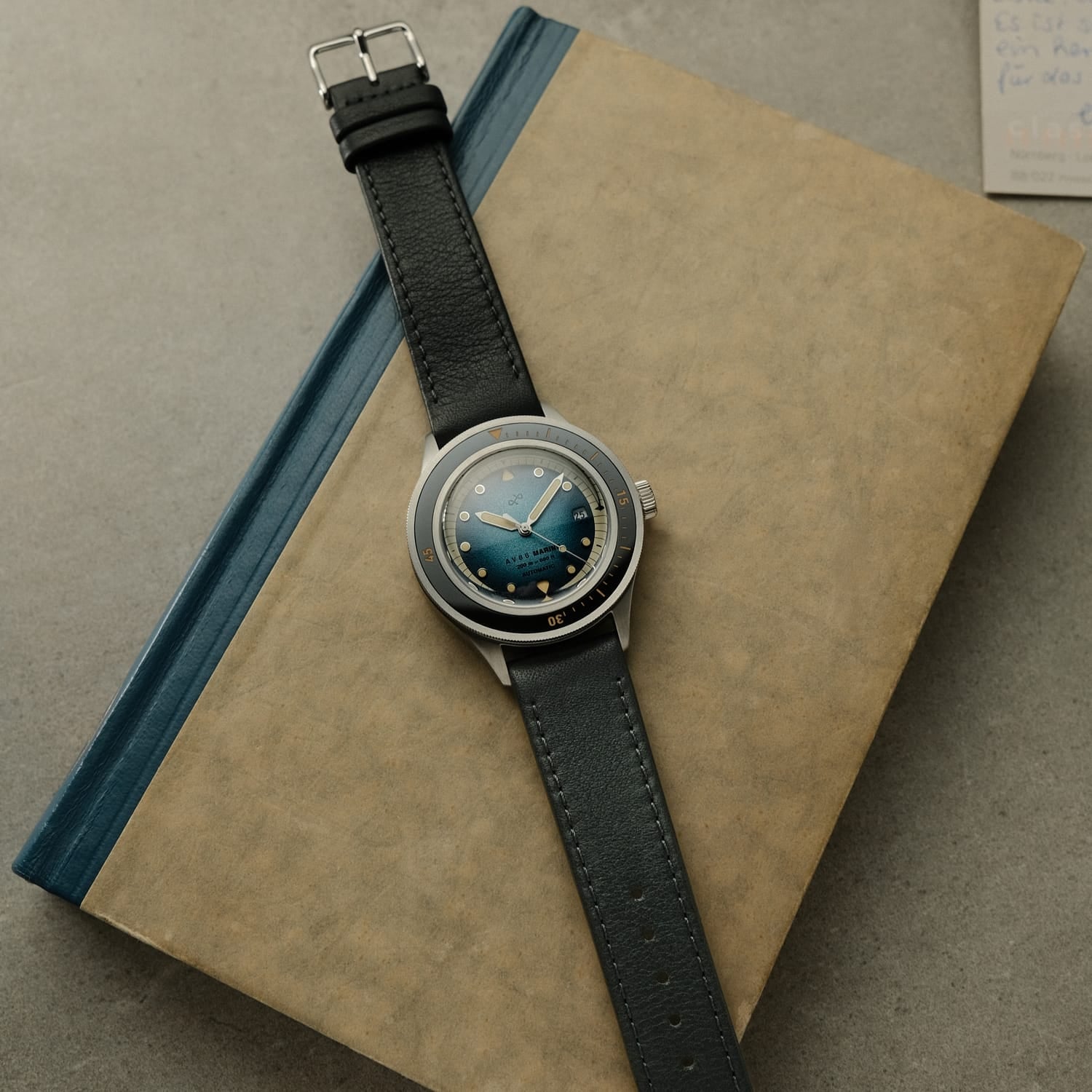 1956 Marine Automatic, Steel / Blue Fade vintage-inspired watch by AV86 with leather strap and blue gradient dial on a book background
