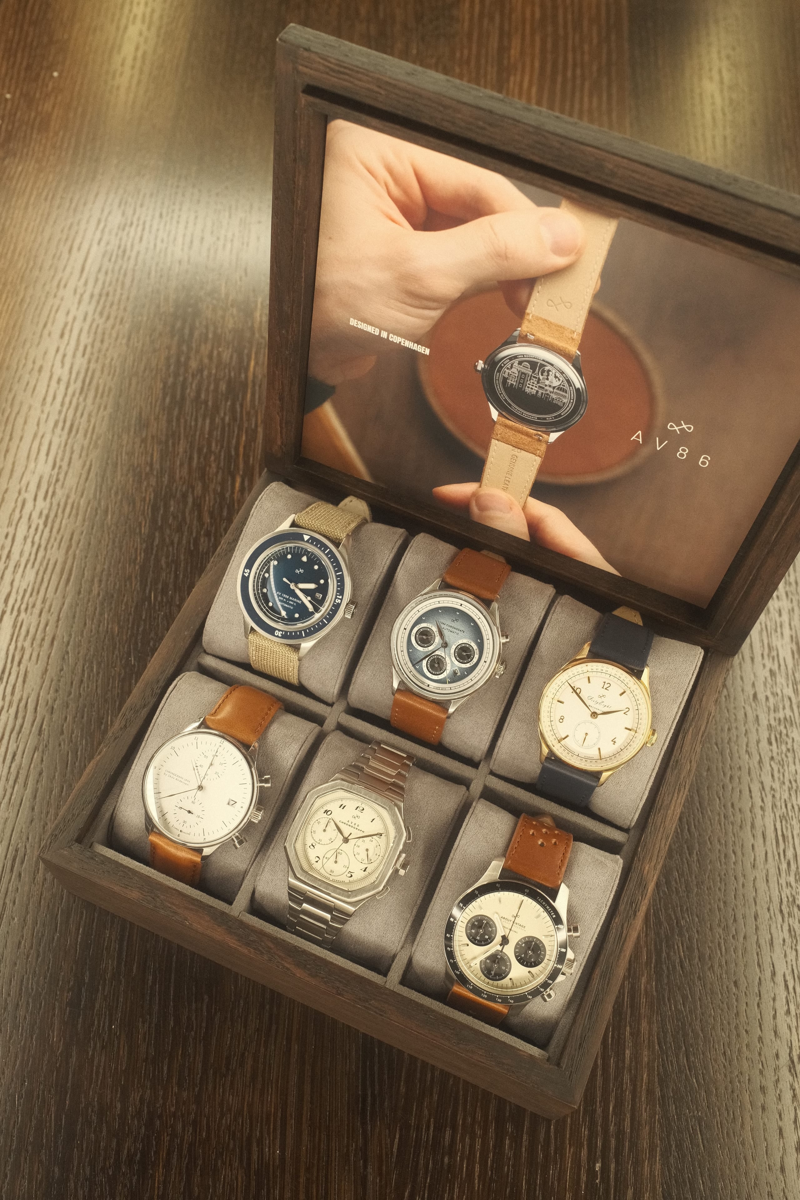 Wooden display box with six diverse wristwatches featuring leather, fabric, and metal straps arranged on soft cushions.