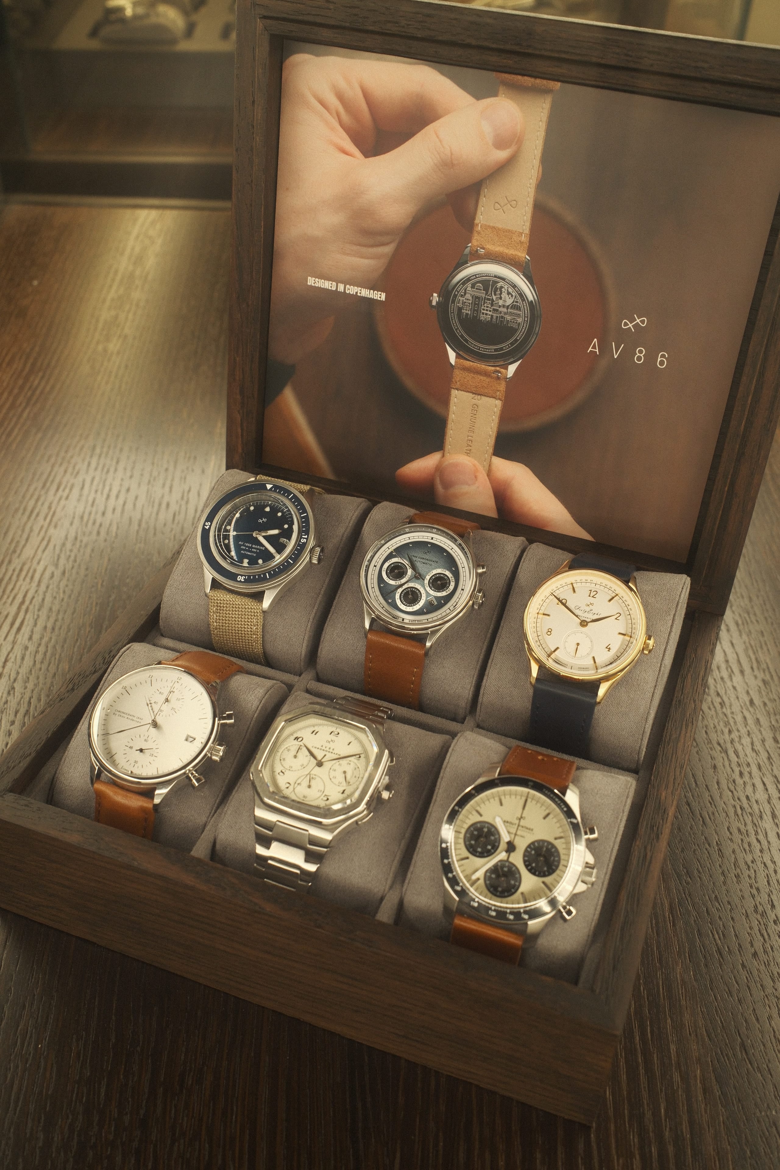 Wooden box with six wristwatches including chronographs, vintage-inspired designs, and leather or metal straps on gray cushions.