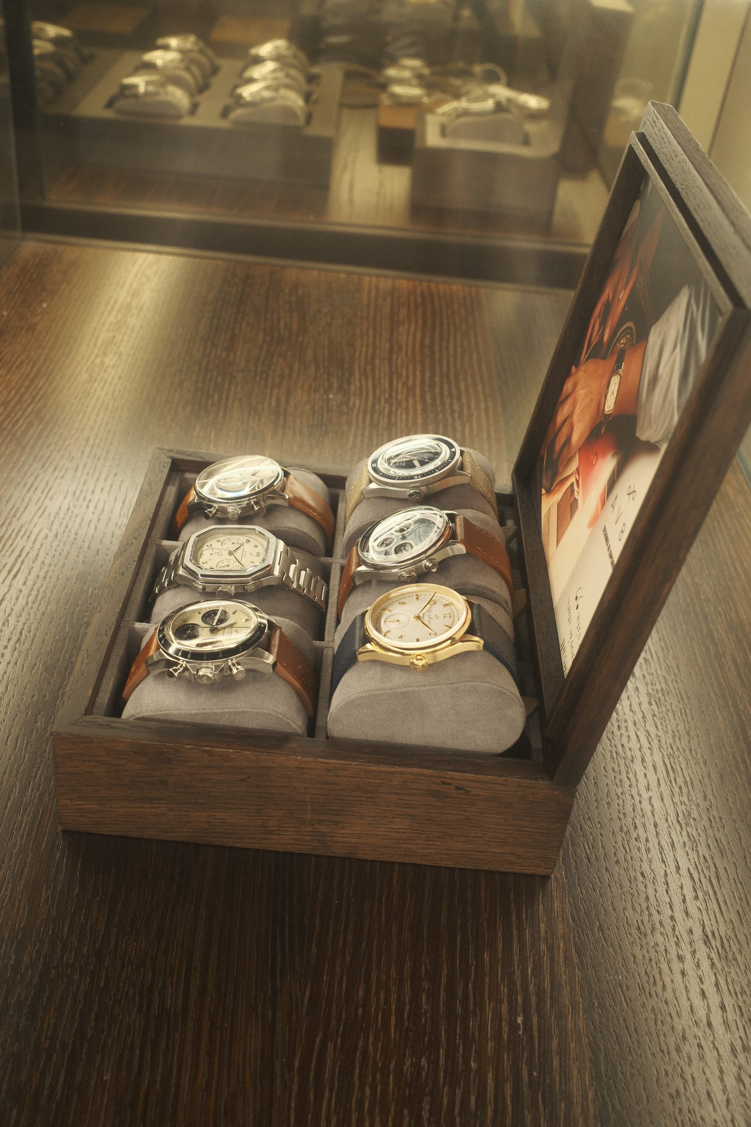 Wooden display box holding six wristwatches with leather and metal straps, arranged on gray cushions beside a framed image.