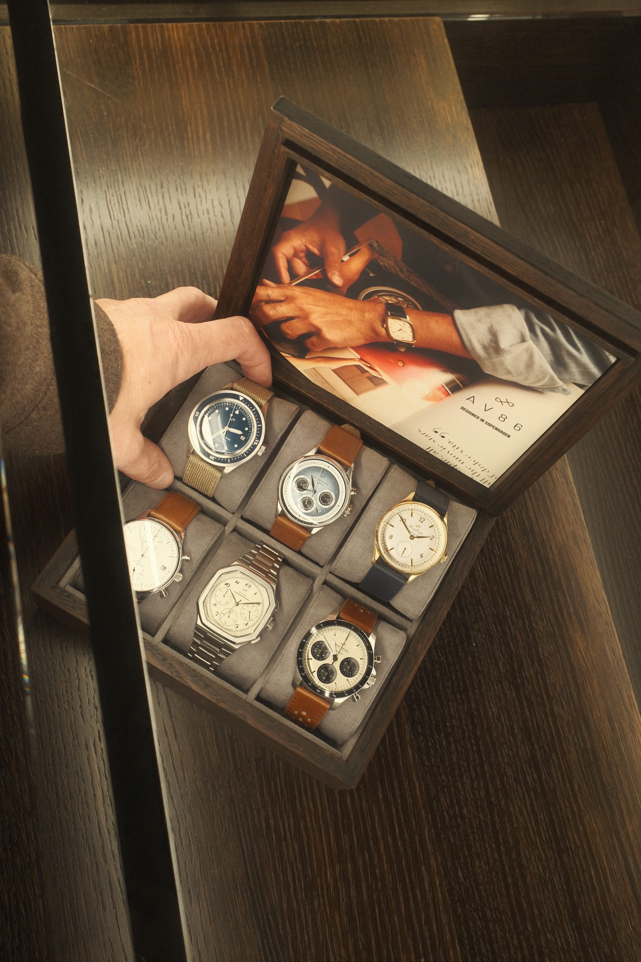 Wooden box holding six wristwatches with diverse designs including metal, leather, and fabric straps on a dark wood surface.