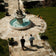 Two men walk along a sunlit stone pathway toward an ornate fountain in a manicured garden, evoking a serene and contemplative atmosphere that complements the refined elegance of the 1564 Chronodate Automatic by AV86.