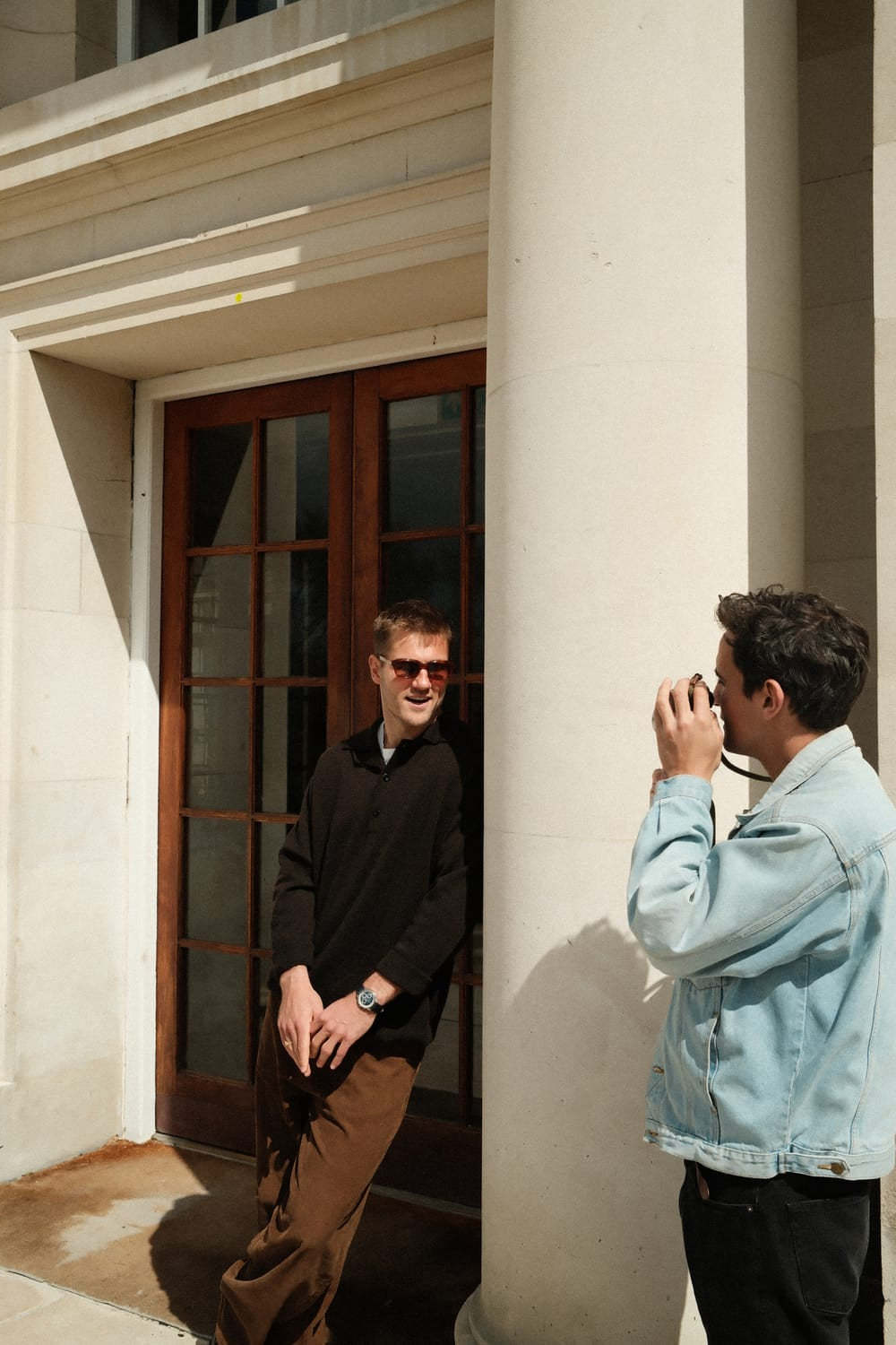 Two men interact beside a sunlit classical building entrance, one wearing sunglasses and a black sweater showcasing the 1564 Chronodate Automatic by AV86, while the other photographs, capturing a moment of casual elegance and Nordic minimalism.