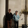 Two men stand casually against a light stone wall in soft sunlight, one wearing brown pants and dark sunglasses, the other in a light denim jacket and black trousers, evoking a relaxed, minimalist Scandinavian style ideal for showcasing the 1564 Chronodate Automatic, Steel / Blue by AV86.