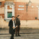 Two men in smart casual attire stand on a cobblestone street by a historic brick building, capturing a relaxed urban mood that complements the refined allure of the 1564 Chronodate Automatic, Steel / Blue by AV86.