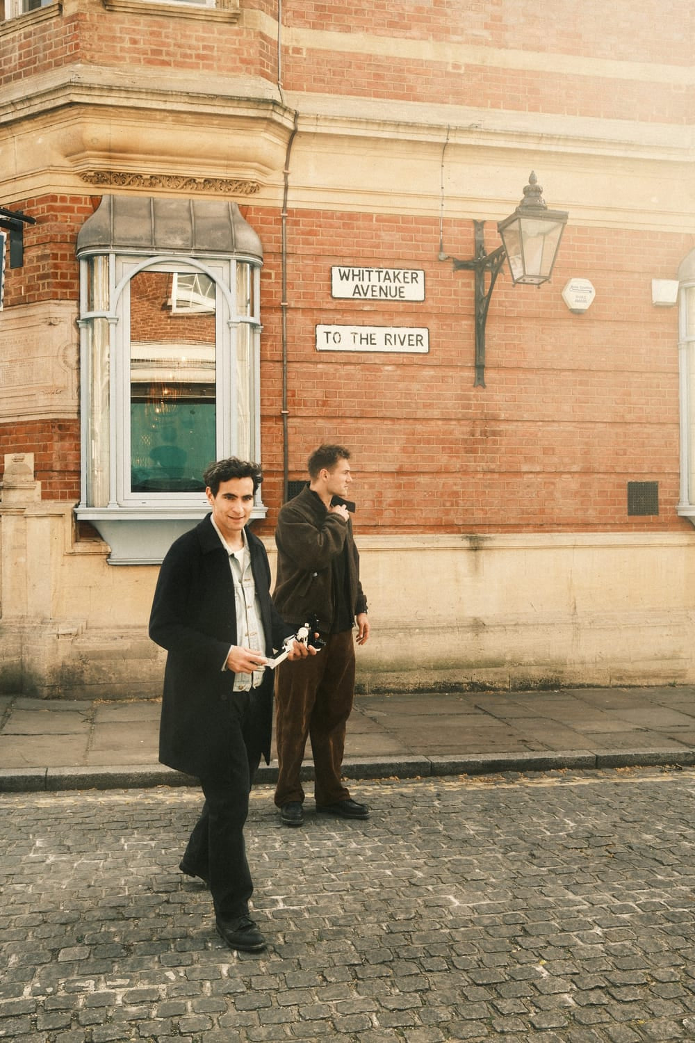 Two men in smart casual attire stand on a cobblestone street by a historic brick building, capturing a relaxed urban mood that complements the refined allure of the 1564 Chronodate Automatic, Steel / Blue by AV86.