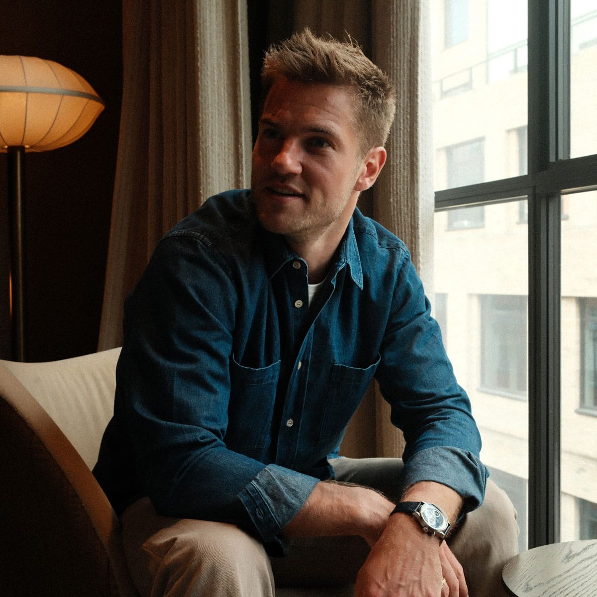 A casually dressed man in a denim shirt sits near a window in soft natural light, creating a relaxed and contemplative Nordic-inspired atmosphere.