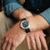 A man dressed in a rolled-up denim shirt and beige trousers casually leans forward, wearing the 1564 Chronodate Automatic, Steel / Blue by AV86 with its polished case and textured blue leather strap, embodying refined Scandinavian style and understated luxury.