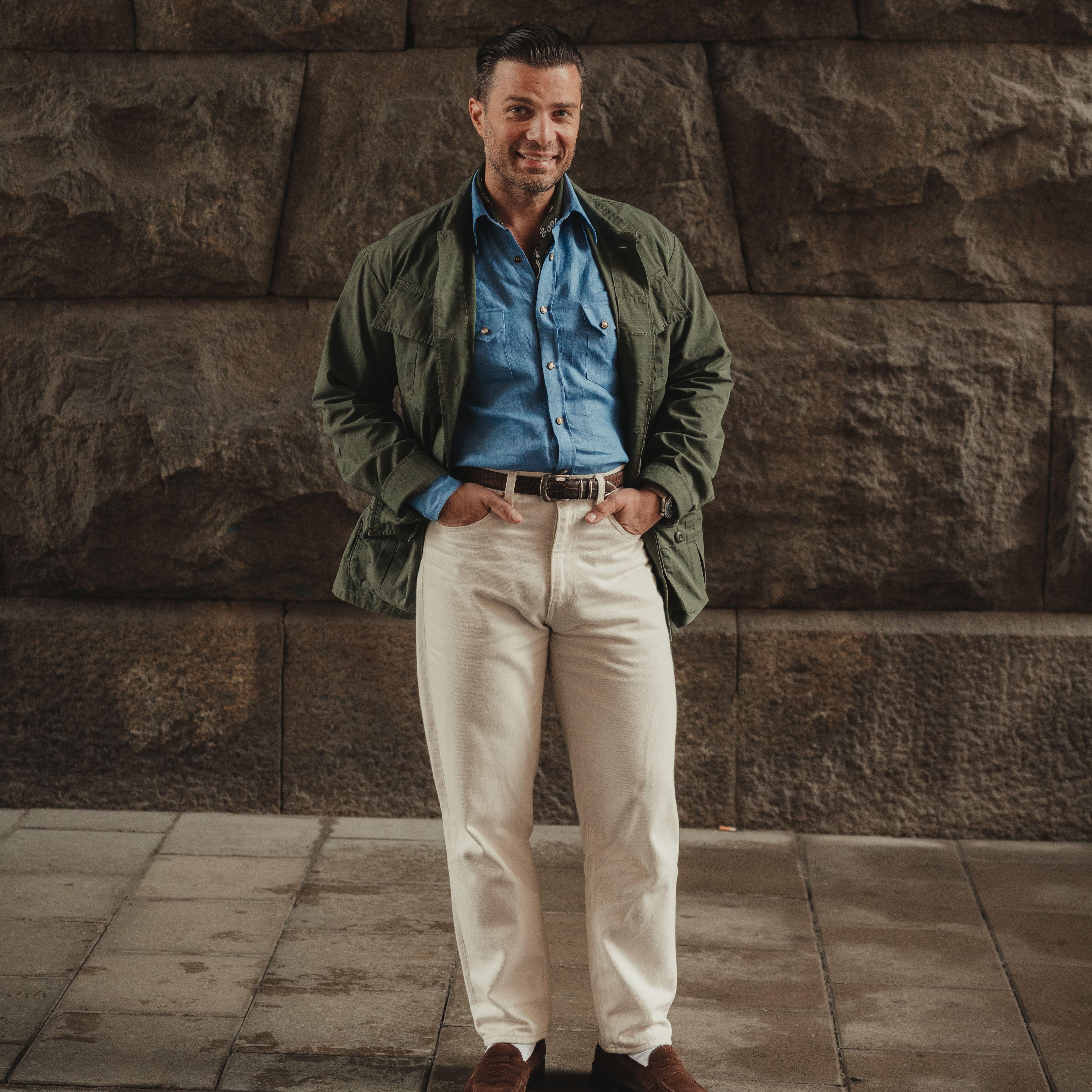 Man in casual outdoor attire with green jacket, blue shirt, beige trousers, and brown shoes against textured stone wall background