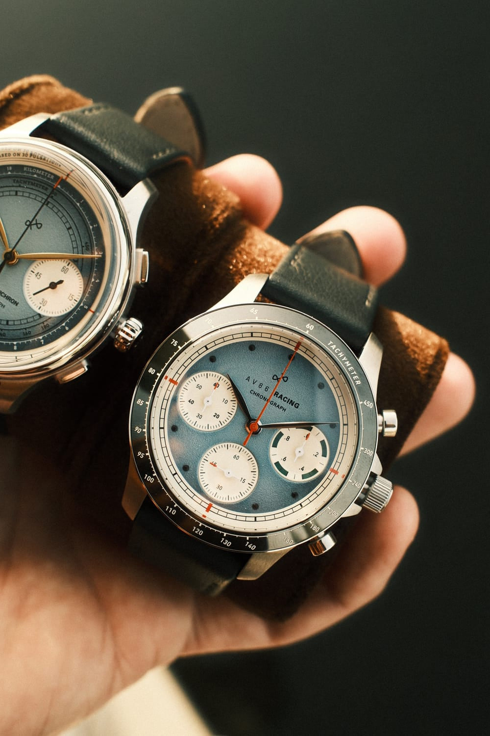 AV86 1960 Racing Chronograph, Steel / Sky Blue & Green quartz watch with tri-compax dial, tachymeter bezel, and leather strap held in hand
