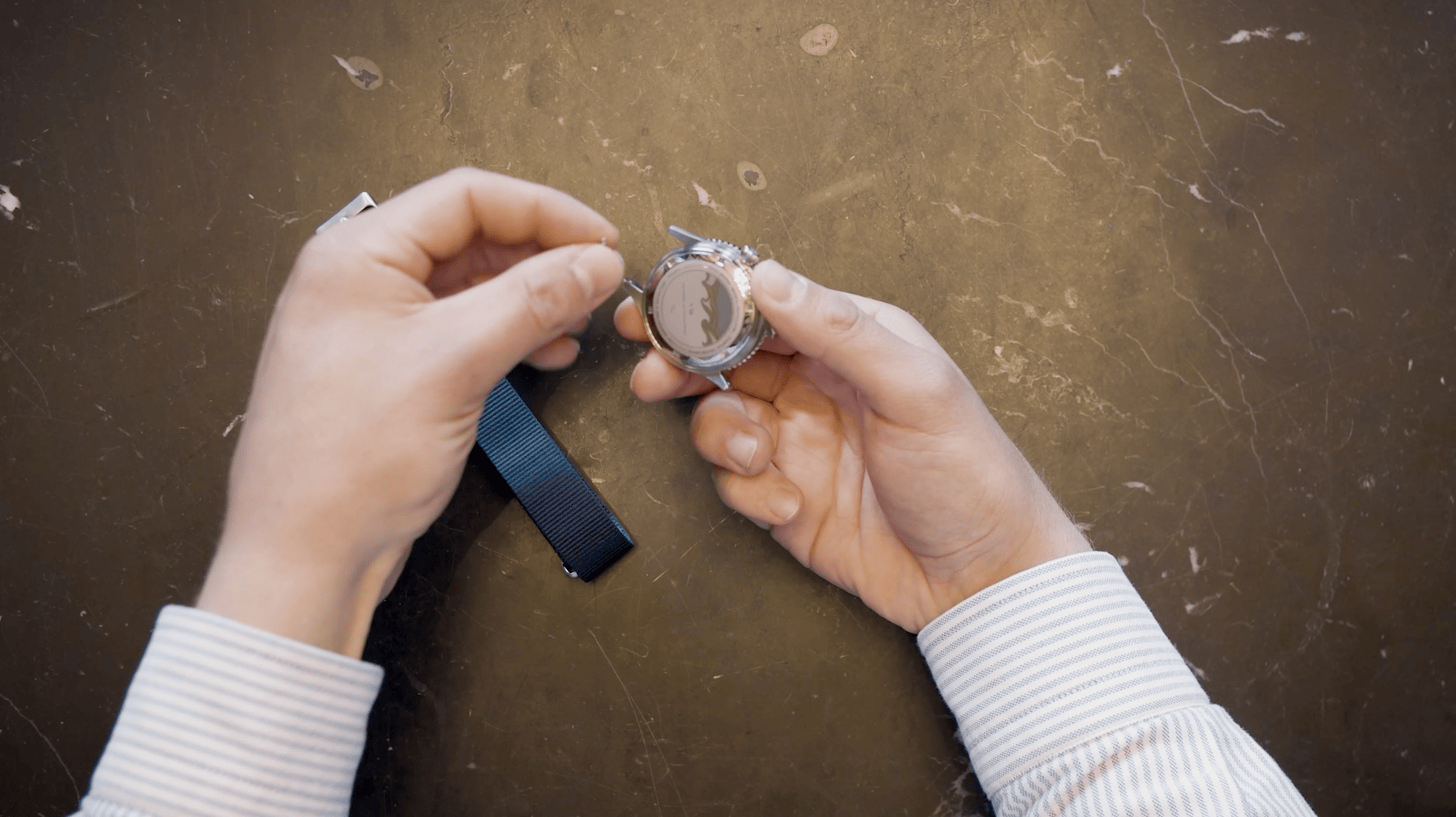 Hands holding a stainless steel watch case while preparing to attach a black NATO fabric strap on a textured brown surface, showcasing detailed craftsmanship.